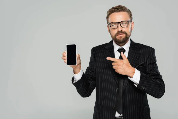 Hombre de negocios barbudo con traje y gafas apuntando al teléfono inteligente con pantalla en blanco aislado en gris - foto de stock