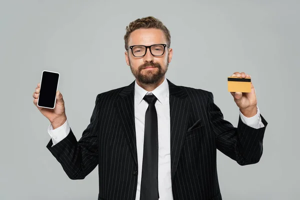 Hombre de negocios barbudo con traje y gafas con teléfono inteligente y tarjeta de crédito aislados en gris - foto de stock
