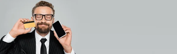 Веселый бизнесмен в костюме и очках, держащий смартфон и кредитную карту изолированными на сером, баннер — стоковое фото