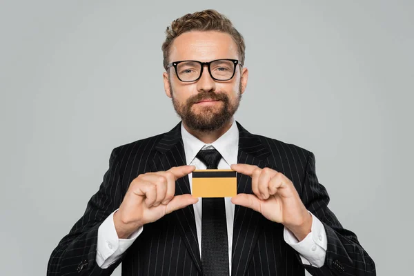 Successful businessman in suit and glasses holding credit card isolated on grey — Stock Photo