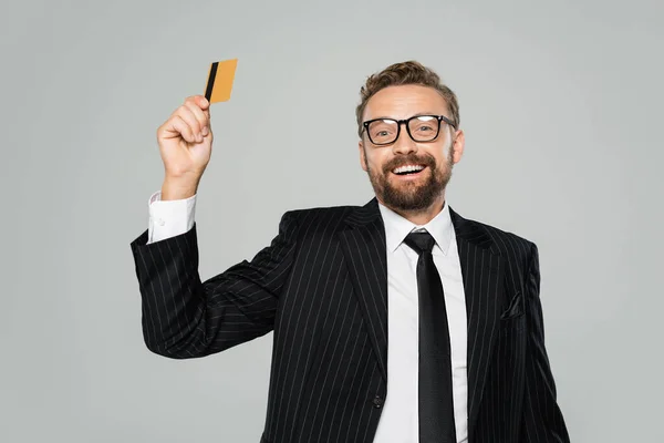 Hombre de negocios positivo con traje y gafas con tarjeta de crédito aislada en gris - foto de stock