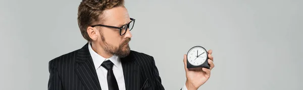 Hombre de negocios barbudo en gafas y ropa formal con reloj despertador vintage aislado en gris, pancarta - foto de stock