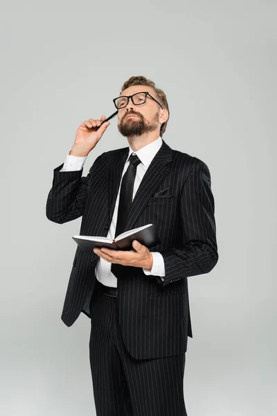 Uomo d'affari pensieroso in occhiali e abbigliamento formale che tiene il quaderno isolato su grigio — Foto stock