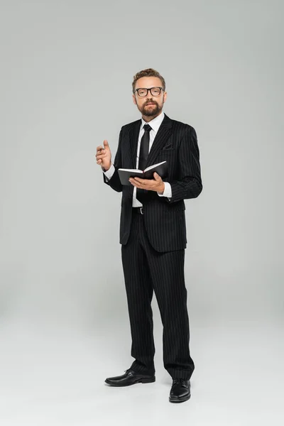 Повна довжина добре одягненого бізнесмена в окуляри і формальний одяг тримає блокнот на сірому — стокове фото