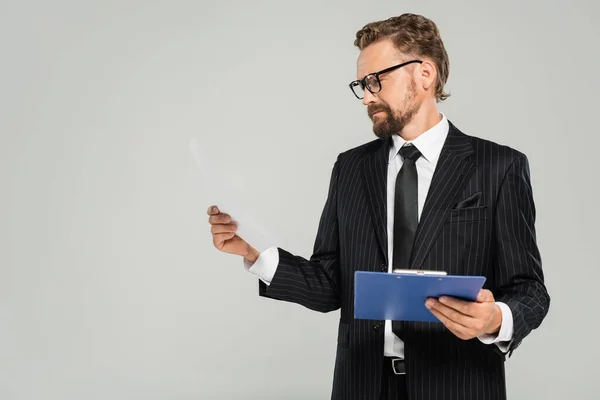 Хорошо одетый бизнесмен в очках держит планшет и смотрит на бумагу, изолированную на сером — стоковое фото