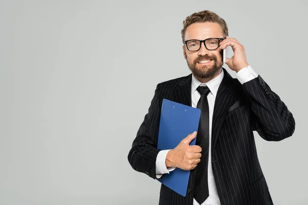 Homme d'affaires heureux dans des lunettes tenant presse-papiers et parlant sur smartphone isolé sur gris — Photo de stock