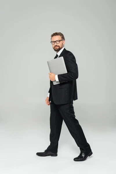 Full length of well dressed businessman in glasses walking with laptop on grey — Stock Photo