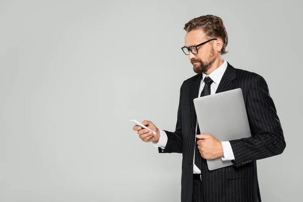 Homme d'affaires bien habillé dans des lunettes tenant ordinateur portable et utilisant smartphone isolé sur gris — Photo de stock