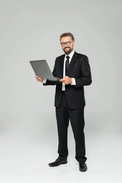 Longitud completa de hombre de negocios feliz y bien vestido en gafas usando el ordenador portátil en gris - foto de stock