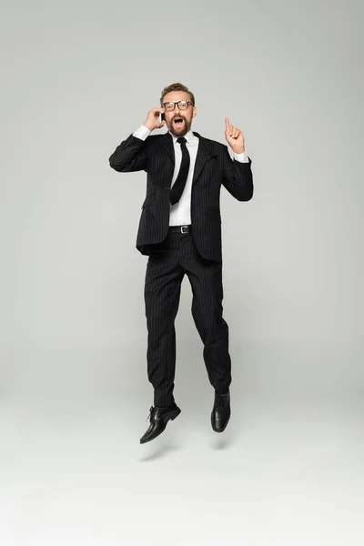 Longitud completa de empresario emocionado en gafas hablando en el teléfono inteligente y apuntando hacia arriba mientras salta en gris - foto de stock