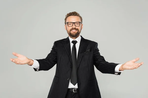 Happy businessman in suit and glasses looking at camera and showing welcoming gesture isolated on grey — Stock Photo