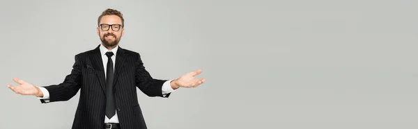 Homme d'affaires heureux en costume et lunettes regardant la caméra et montrant geste accueillant isolé sur gris, bannière — Photo de stock