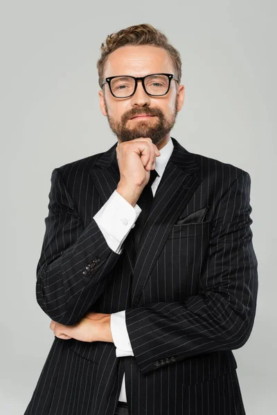 Hombre de negocios barbudo con traje y gafas tomados de la mano cerca de la barbilla mirando a la cámara aislada en gris - foto de stock