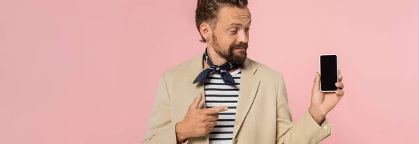 Бородатый французский мужчина в блейзере и шарф на шее указывая на смартфон с чистым экраном изолированы на розовый, баннер — стоковое фото