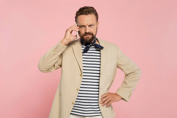 Hombre francés disgustado en chaqueta y cuello bufanda hablando en el teléfono inteligente, mientras que de pie con la mano en la cadera aislado en rosa - foto de stock