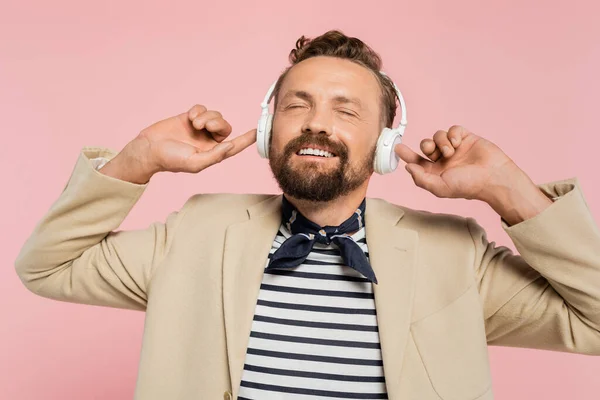 Hombre francés satisfecho en chaqueta y cuello bufanda escuchar música en auriculares inalámbricos aislados en rosa - foto de stock