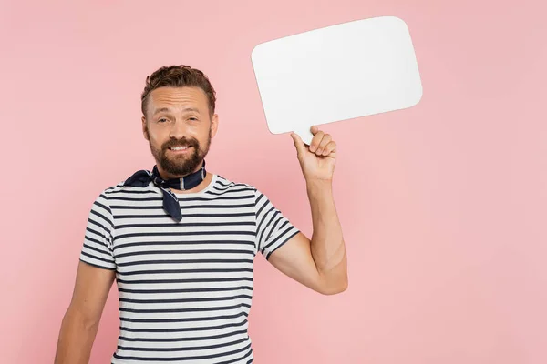 Happy french guy in striped t-shirt and neck scarf holding speech bubble isolated on pink — Stock Photo