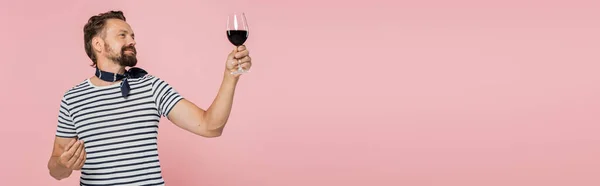 Pleased man in striped t-shirt holding glass of red french wine isolated on pink, banner — Stock Photo
