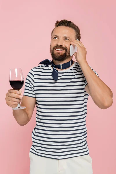 Fröhlicher Mann im gestreiften T-Shirt, der ein Glas französischen Rotwein in der Hand hält und isoliert auf einem Smartphone in rosa spricht — Stockfoto