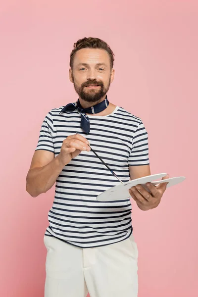 Cheerful french artist with neck scarf holding palette and paintbrush isolated on pink — Stock Photo