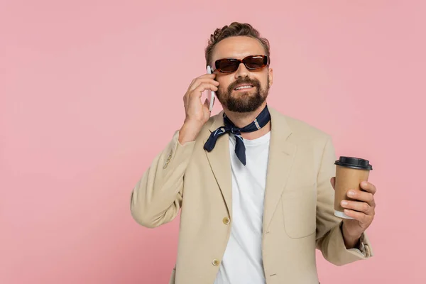 Hombre francés feliz en la bufanda del cuello y gafas de sol sosteniendo café para ir y hablar en el teléfono inteligente aislado en rosa - foto de stock