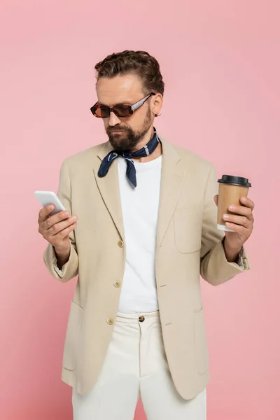 Elegante hombre francés en la bufanda del cuello y gafas de sol sosteniendo café para ir y el uso de teléfono inteligente aislado en rosa - foto de stock
