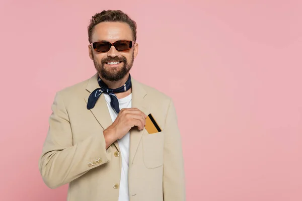 Щасливий чоловік в стильних сонцезахисних окулярах і шарфі шиї кладе кредитну картку в кишеню на блістері ізольовано на рожевому — стокове фото