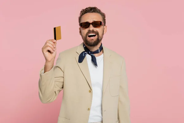 Excited man in stylish sunglasses and neck scarf holding credit card isolated on pink — Stock Photo