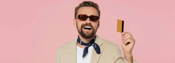 Uomo eccitato in eleganti occhiali da sole e sciarpa collo con carta di credito isolata su rosa, banner — Foto stock