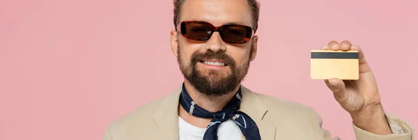 Uomo felicissimo in occhiali da sole alla moda e sciarpa collo con carta di credito isolata su rosa, banner — Foto stock