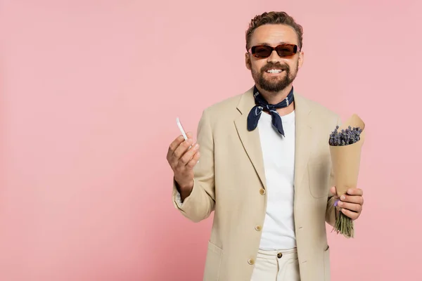 Überglücklicher Mann in stylischer Sonnenbrille und Halstuch mit Blumen in Papierfolie und Smartphone isoliert auf rosa — Stockfoto