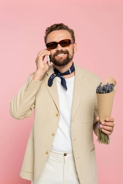 Glücklicher Mann mit stylischer Sonnenbrille und Halstuch, der Blumen hält und auf dem Smartphone isoliert auf rosa spricht — Stockfoto