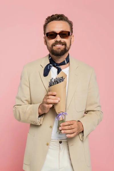 Восторженный человек в стильных солнцезащитных очках и шарф с цветами в бумажной обертке изолированы на розовый — стоковое фото