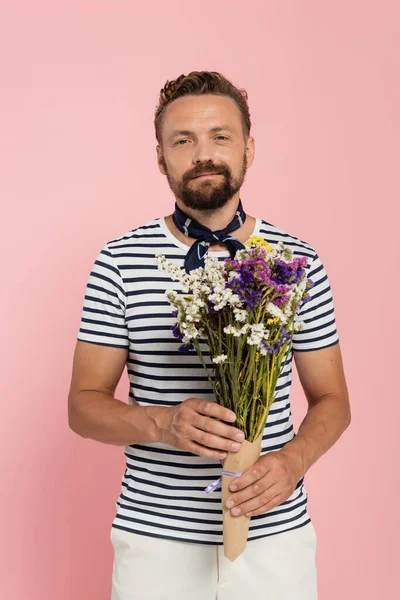 Felice uomo in t-shirt a righe e sciarpa collo che tiene fiori in involucro di carta isolato su rosa — Foto stock