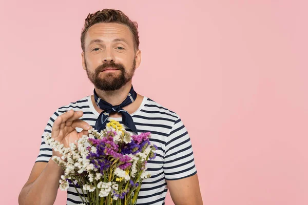Uomo allegro in t-shirt a righe e sciarpa collo tenendo fiori selvatici in involucro di carta isolato su rosa — Foto stock