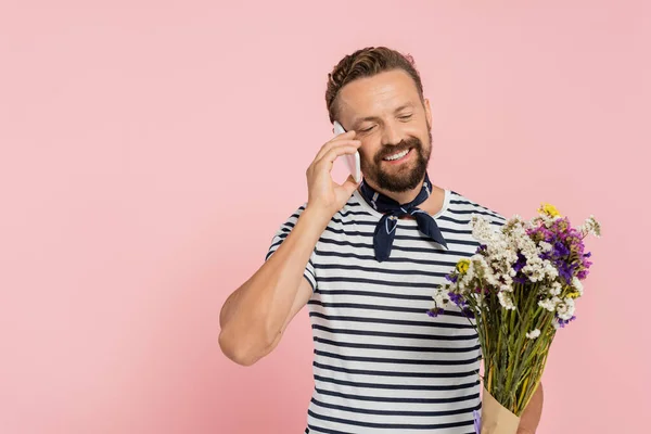 Fröhlicher Mann in gestreiftem T-Shirt und Halstuch, der Wildblumen in der Hand hält und isoliert auf einem Smartphone in rosa spricht — Stockfoto