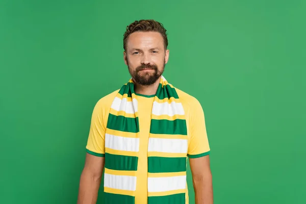 Бородатый спортивный болельщик в полосатом шарфе и желтой футболке, смотрящий на камеру, изолированную на зеленом — стоковое фото