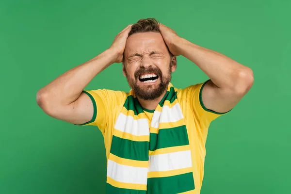 Емоційний спортивний фанат у смугастому шарфі та жовтій футболці, що тягне волосся ізольовано на зеленому — стокове фото
