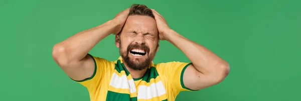 Эмоциональный фанат спорта в полосатом шарфе и желтой футболке дергая его волосы изолированы на зеленый, баннер — стоковое фото
