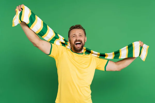 Збуджений спортивний вентилятор у жовтій футболці, що тримає смугастий шарф і дивиться на камеру ізольовано на зеленому — стокове фото