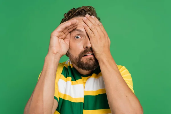 Tifoso spaventato in sciarpa a righe e t-shirt gialla che copre il viso isolato sul verde — Foto stock