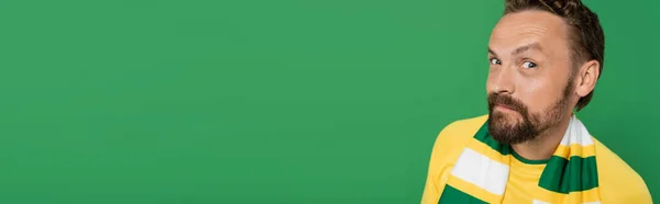 Fan de sport suspect en écharpe rayée et t-shirt jaune regardant la caméra isolée sur vert, bannière — Photo de stock