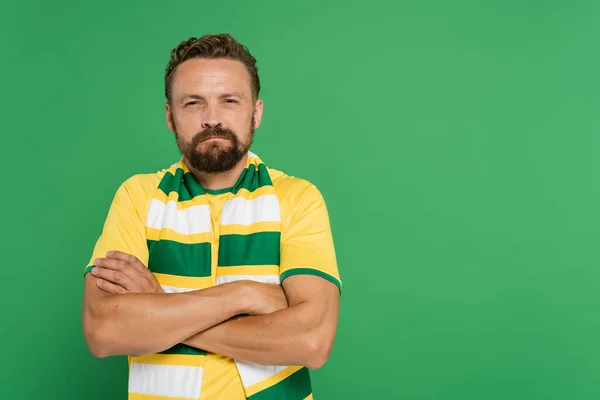 Éventail de football offensé en écharpe rayée et t-shirt jaune debout avec les bras croisés isolés sur vert — Photo de stock