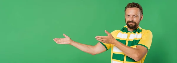 Abanico de fútbol barbudo en bufanda a rayas y camiseta amarilla mirando a la cámara mientras señala con las manos aisladas en verde, pancarta - foto de stock