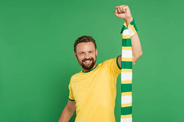 Веселий футбольний вентилятор у жовтій футболці, що тримає смугастий шарф і дивиться на камеру ізольовано на зеленому — стокове фото