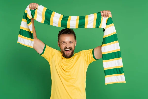 Emocionado fanático del fútbol en camiseta amarilla sosteniendo bufanda a rayas y mirando a la cámara aislada en verde - foto de stock