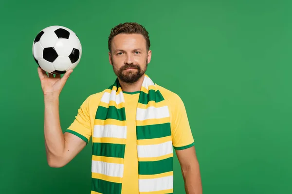 Abanico de fútbol barbudo en bufanda a rayas y camiseta amarilla que sostiene el fútbol aislado en verde - foto de stock