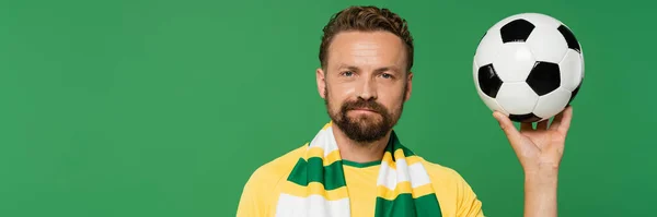 Bearded sports fan in striped scarf and yellow t-shirt holding football isolated on green, banner — Stock Photo