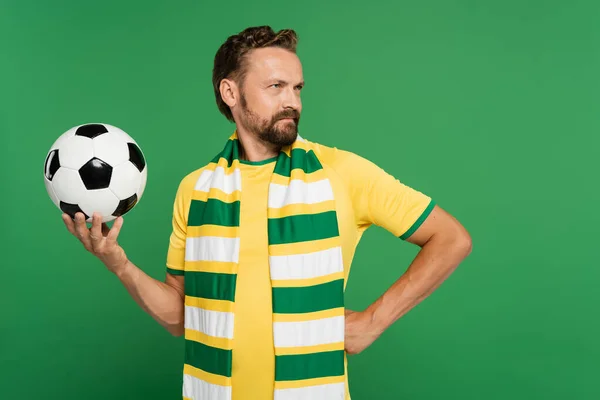 Abanico de fútbol barbudo en bufanda a rayas y camiseta amarilla que sostiene el fútbol mientras está de pie con la mano en la cadera aislado en verde - foto de stock