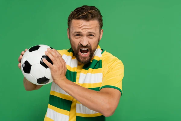 Emotional soccer fan in striped scarf and yellow t-shirt holding football isolated on green — Stock Photo
